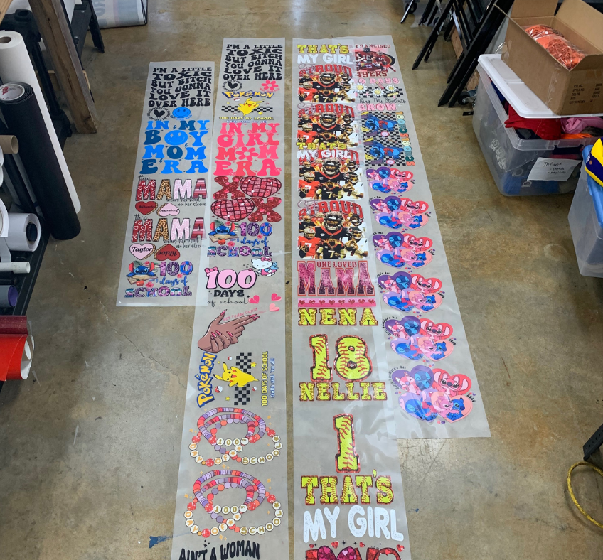 Custom Direct to Film (DTF) gang sheets laid out on a table, featuring an array of designs for t-shirts including logos, graphics, and text, ready for heat transfer application.