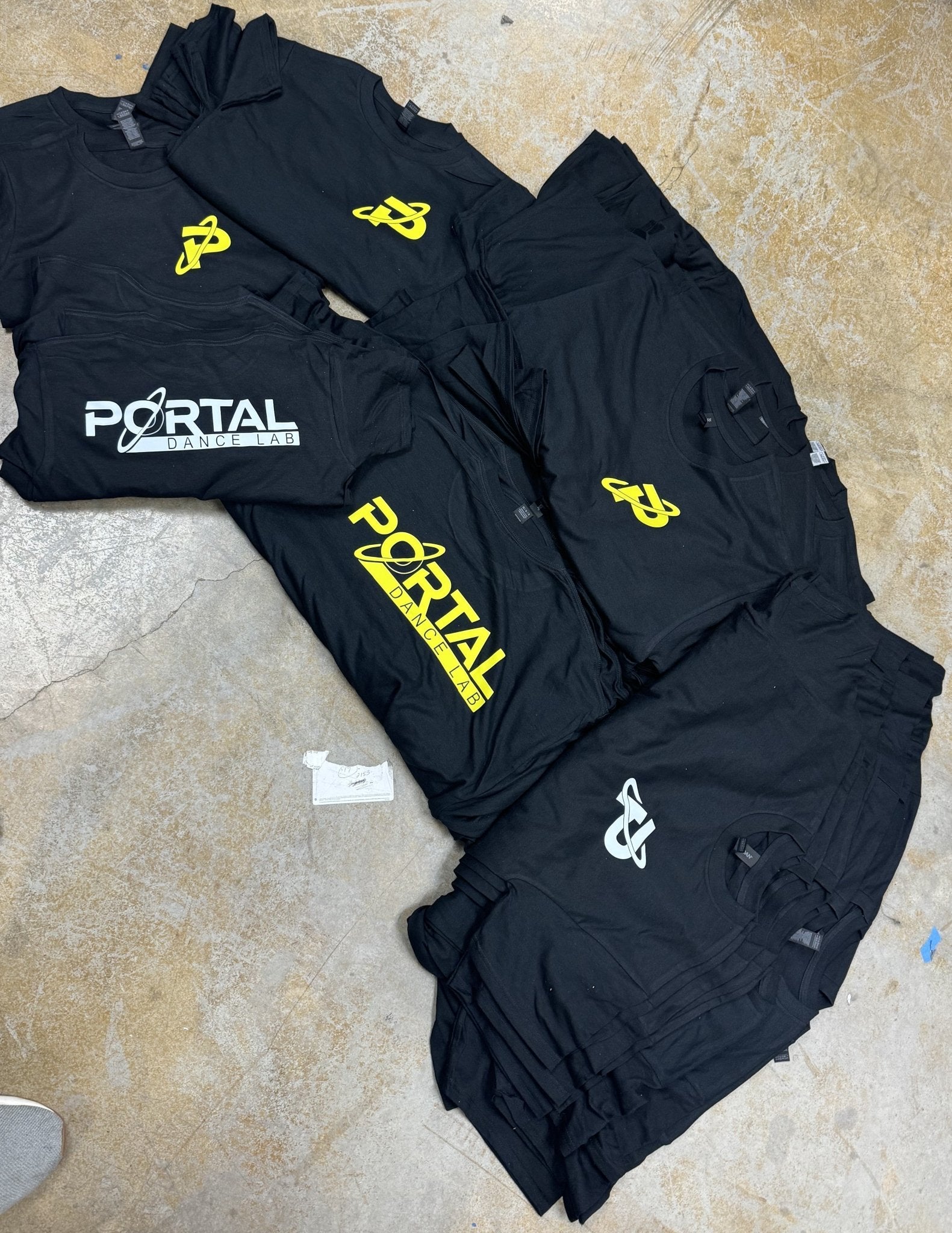 Black Custom Gildan Youth Heavy Cotton™ T-Shirts by Gildan, featuring yellow and white "Portal Dance Lab" logos, are arranged on a concrete floor. Personalize this soft, eco-friendly tee with your own logo for added style.